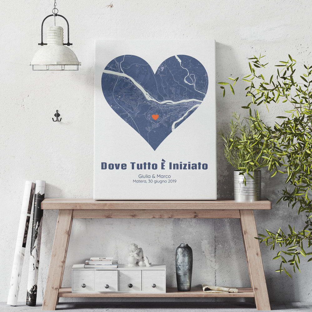 Stampa con mappa a cuore e segnaposto arancione su una consolle in legno, tra una lampada e una pianta